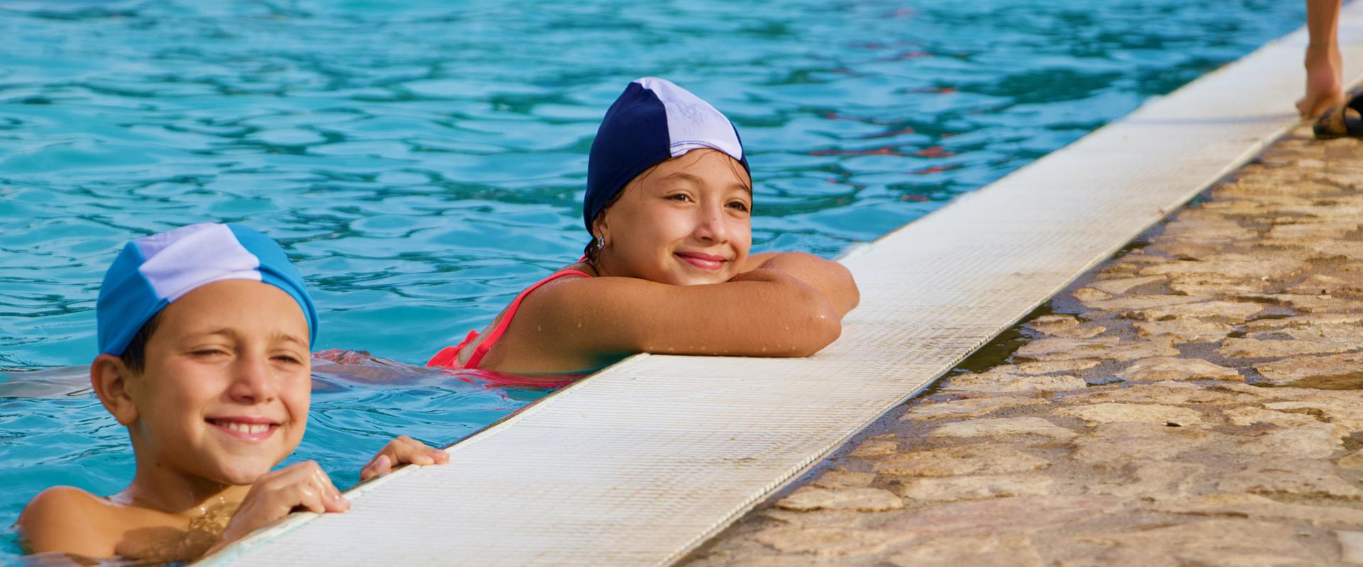 Bambini con cuffie da nuoto saltano in piscina, mentre altri si rilassano sui lettini.