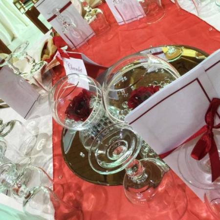 Tavola apparecchiata con bicchieri, tovaglioli rossi e decorazioni floreali su specchio.