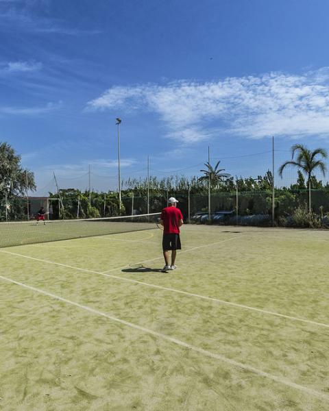 Partita di tennis su campo all'aperto, con cielo sereno e vegetazione circostante.