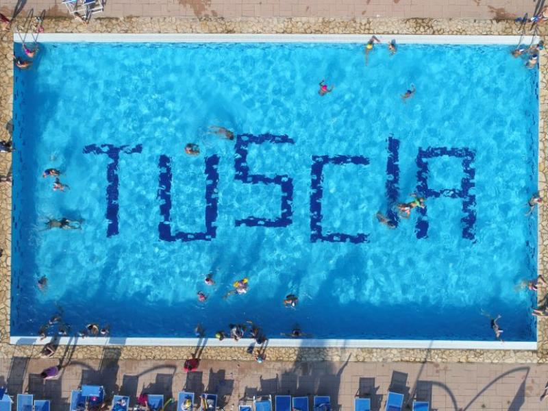 Piscina con la scritta 'TUSCIA' formata da persone in acqua.